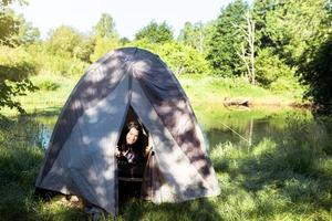 en glad kvinna i rutig skjorta tittar ut ur ett turisttält på en vandring på flodstranden på morgonen. camping i naturen, övernattning i det vilda, familjesemester och äventyr. foto