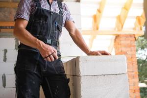 byggnadsarbetare på byggarbetsplatsen mäter längden på fönsteröppningen och tegelväggen med måttband. stugan är gjord av porösa betongblock, arbetskläder - jumpsuit och baseballkeps foto