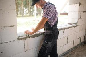 byggnadsarbetare på byggarbetsplatsen mäter längden på fönsteröppningen och tegelväggen med måttband. stugan är gjord av porösa betongblock, arbetskläder - jumpsuit och baseballkeps foto