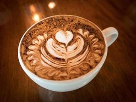 cappuccino med färskt mjölkskum och lattekonst i ett vitt keramiskt glas på ett träbord på ett kafé. foto