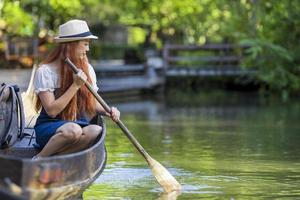 ung asiatisk kvinna resenären roddar träbåt med paddel under sin resa till Thailand och besöker den lokala flytande marknaden med lugn tropisk natur i Sydostasien foto