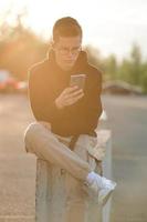 en snygg ung kille i luvtröja, lediga byxor och vita sneakers. en kille med hörlurar sitter och använder en smartphone. foto