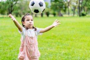 leende liten flicka stående kasta fotboll på grön fotbollsplan i sommardag. porträtt av liten flicka idrottsman leker med en boll på stadion. aktiv barndom koncept. kopieringsutrymme foto