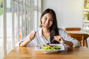 porträtt av attraktiv asiatisk leende ung kvinna som äter sallad i restaurangen, en ung glad tjej som äter en hälsosam lunch foto