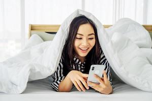 glad ganska ung asiatisk kvinna som använder mobiltelefon under filten i sängen i heminredning, spelar online mobilspel, tittar på roliga videor, njuter av tidsfördriv hemma. foto