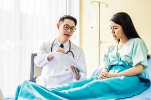 manlig läkare pratar med kvinnlig patient i sjukhussäng, medan han pekar anteckningar på urklipp, medicin och hälsovårdskoncept, selektiv fokuspunkt foto