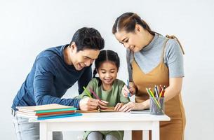 Asiatisk ung mor och far med lilla dotter sitter vid skrivbordet lärande och skrivande i bok med penna göra läxor hemma. utbildning koncept. foto