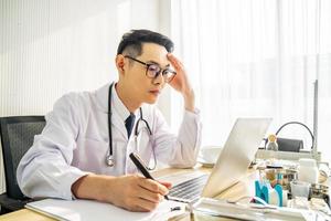 orolig asiatisk man läkare som arbetar på datorn vid skrivbordet på sjukhuskliniken, orolig, tänker hårt, hälsokoncept för covid19. foto