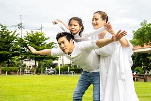 porträtt av asiatisk lycklig familj på tre som har roligt tillsammans i parken, lilla dotter sitter på pappas rygg och mamma ler går och har roliga stunder bra tid i sommarparken hemma foto