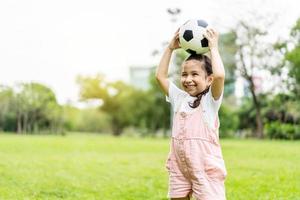 leende liten flicka står och håller fotbollen på grön fotbollsplan i sommardag. porträtt av liten flicka idrottsman leker med en boll på stadion. aktiv barndom koncept. kopieringsutrymme foto