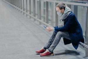 trött turist tar paus, använder mobiltelefon, klädd i jacka, jeans och sneakers, bär skyddsmask mot coronavirus, bryr sig om hälsan på allmän plats, läser information. säkerhetskoncept foto