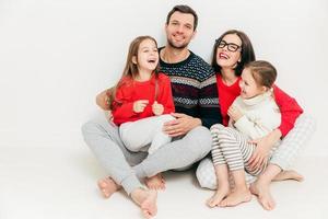 studiobild av glada familjemedlemmar som sitter på golvet med bara fötter, omfamnar varandra, har glada uttryck, skratta när du hör positiva nyheter eller rolig historia, isolerad på vit bakgrund. föräldrar, barn foto