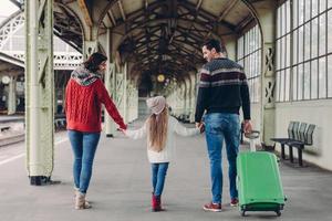 horisontell vy av tillgivna föräldrar och deras dotter håller varandra i händerna, ska resa utomlands, posera på järnvägsstationen, bära grön väska, se glad ut, kommunicera. familjeresa foto