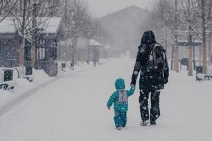 bakifrån av tillgiven far och litet manligt barn håller hand, gå längs snöig gata under kraftigt snöfall på vintern, njuta av rekreationstid, gå hem, klädd i varma kläder, bära ryggsäck foto