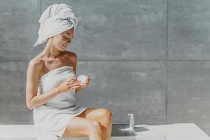 horisontell bild av avslappnad vacker kvinna applicerar bodylotion och närande kräm efter duschen, poserar i badrummet mot en grå vägg, sitter inlindad i handduk, genomgår skönhetsbehandlingar. spa koncept foto
