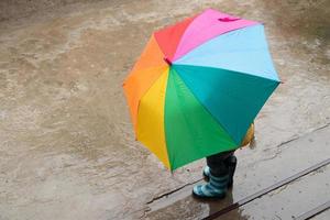 en 3-årig flicka gömmer sig i regnet under ett färgat paraply foto