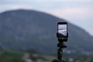 smartphonekamerastativ på ett stativ och fotograferar en pittoresk kvällssolnedgång över det tropiska havet och bergen. foto
