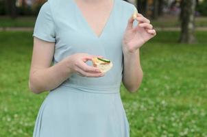 en ung flicka håller en smörgås med skinka, ost och en skiva gurka i handen på en picknick i parken. hälsosam sommarpicknick i trädgården eller parken. foto