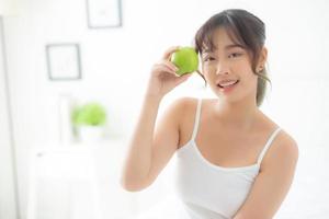 vacker ung asiatisk kvinna som håller och äter grönt äpple frukt i sovrummet hemma, livsstil för näring flicka hälsosam och omsorg viktminskning, hälsa och välbefinnande koncept. foto