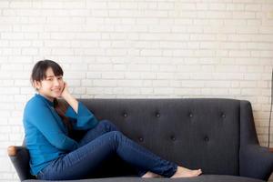 vackert porträtt ung asiatisk kvinna sitter och ler glad och tittar på kameran på soffan med casual i vardagsrummet, flicka glad och koppla av på soffan hemma, livsstilskoncept. foto