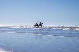 man och kvinna rider hästar längs kustlinjen på stranden mot klar himmel foto