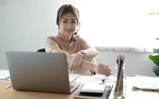 Asiatisk affärskvinna pratar med kollega team i videosamtal konferens skriver anteckning på bok med leende ansikte. kvinna som använder bärbar dator och hörlurar för onlinemöte. foto
