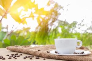 vit kaffekopp med glasunderlägg och kaffebönor placerade på brun säckväv på naturlig bakgrund. foto