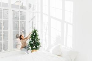 vintersemester, nyårskoncept. glad ung brunett kvinna har festlig stämning, dekorerar julgranen hemma, förbereder sig för firande, poserar i mysigt sovrum med vita väggar på fönsterbrädan. foto