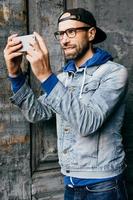 porträtt av snygg kille med skägg som bär trendiga kläder som håller mobiltelefon och gör selfie som är nöjd och ler av glädje in i kameran. avslappnad man med attraktivt utseende poserar i kameran foto