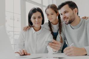 glad vänlig familj av far, mor och dotter sitter framför datorn, kontrollera saldo på kreditkort, gör shopping online, köp något nödvändigt, använd höghastighetsanslutning till internet. foto