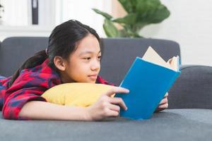 söt liten flicka i fritidskläder läser en bok liggande på en soffa i vardagsrummet foto