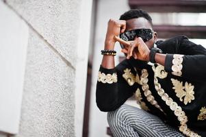 mega snygg afrikansk man i traditionell jacka pose. fashionabla svart kille i solglasögon med cigarr. foto