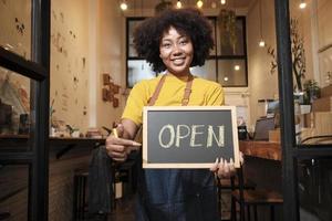 en afrikansk amerikansk kvinnlig startbarista står vid casual cafédörren, tittar på kameran och visar öppen skylt, glada och glada leenden med jobb i kaféservice och nya företagare. foto