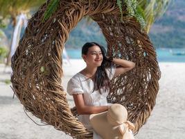 attraktiv asiatisk kvinna sitta på oval trä boet på vit sandstrand tittar på havsutsikt och leende foto