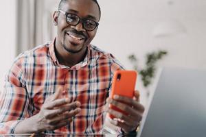 glad afrikansk amerikansk man som bär glasögon som håller smartphone använder moderna appar som sitter vid laptop foto