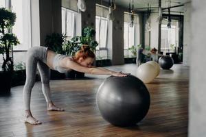 smal kvinna som sträcker sig tillbaka med träningsboll medan du tränar i modern fitnessstudio foto