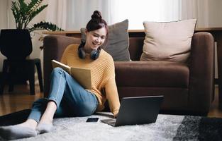 unga asiatiska kvinnor som lyssnar på musik i hörlurar attraktiv asiatisk tjej tittar på den bärbara datorn som sitter i vardagsrummet och spenderar fritid hemma foto