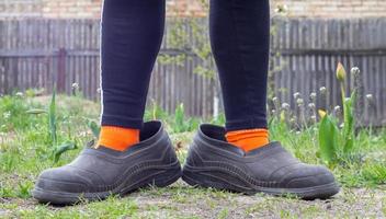 kvinnors ben i överdimensionerade gummigaloscher mot landsbygdens bakgrund. ben i svarta leggings, orange strumpor och smutsiga galoscher i ett förortsområde. väldigt roligt foto. foto