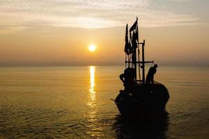 siluett av två personer på en fiskebåt som är på väg att fiska i morgonsolen. foto