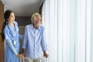 Hospicesjuksköterska stöder kaukasisk man att använda rollator medan han tittar ut genom fönstret på pensionärshem för rehabilitering av hemtjänst och återhämtning efter behandling foto