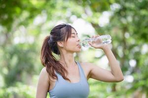 idrottsman ung vacker kvinna dricker vatten från en plastflaska i sommarens gröna park, sportkvinna som dricker vatten efter träningen foto