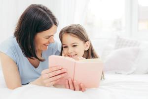 inomhusbild av ett trevligt kvinnligt barn med ett nyfiket uttryck, läser intressant bok tillsammans med sin tillgivna mamma, tittar på färgglada bilder, ligger på sängen i ett rymligt sovrum. barndom foto