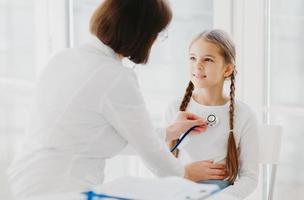 omtänksam kvinnlig barnläkare lyssnar barnets lungor eller hjärta med phonendoscope. liten flicka kommer på undersökning på privat klinik, har läkarbesök varje år, förklarar symtom på sin sjukdom foto