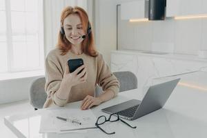 leende kvinnlig kontorsarbetare i headset med smartphone medan du sitter på sin arbetsplats med laptop foto