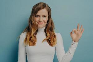 glad ung europeisk kvinna som visar okej gest isolerad över pastellblå studiovägg foto
