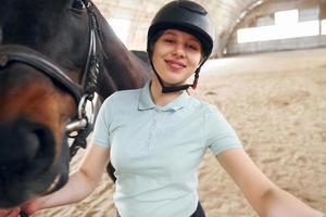 gör en härlig selfie. en ung kvinna i jockeykläder förbereder sig för en tur med en häst på ett stall foto
