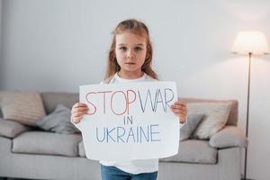 liten flicka står hemma och håller banderoll med stop war i ukrainsk text foto