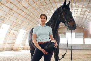 står och håller skyddshatt i händerna. en ung kvinna i jockeykläder förbereder sig för en tur med en häst på ett stall foto