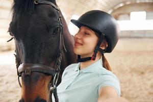 gör en härlig selfie. en ung kvinna i jockeykläder förbereder sig för en tur med en häst på ett stall foto