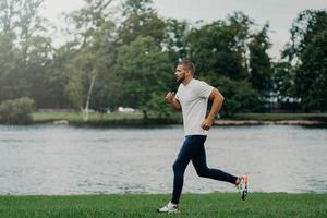 atlet skäggig man springer längs vackert landskap, visar uthållighet, klädd i aktivt slitage, joggar nära floden, andas frisk luft. sport, träning, fitness och träningskoncept foto